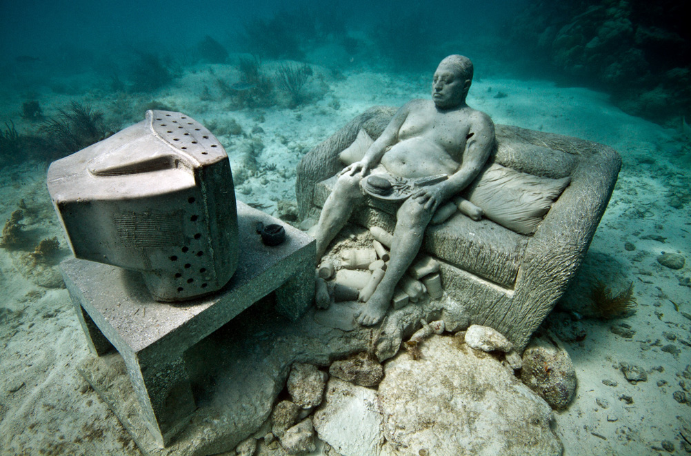The Inertia sculpture designed by Jason deCaires Taylor in Punta Nizuc&#039;s Underwater Sculpture Park in Mexico depicts an man seating on a sofa watching TV while eating a hamburger