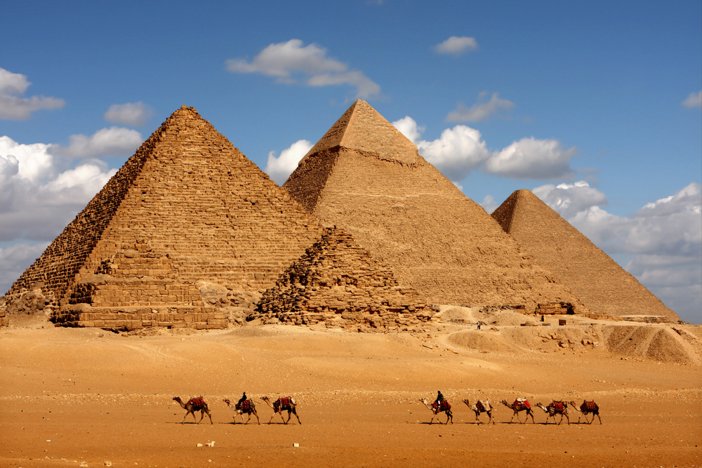 A group of camels with their riders approach the Pyramids of Giza in Egypt on a partly cloudy day.