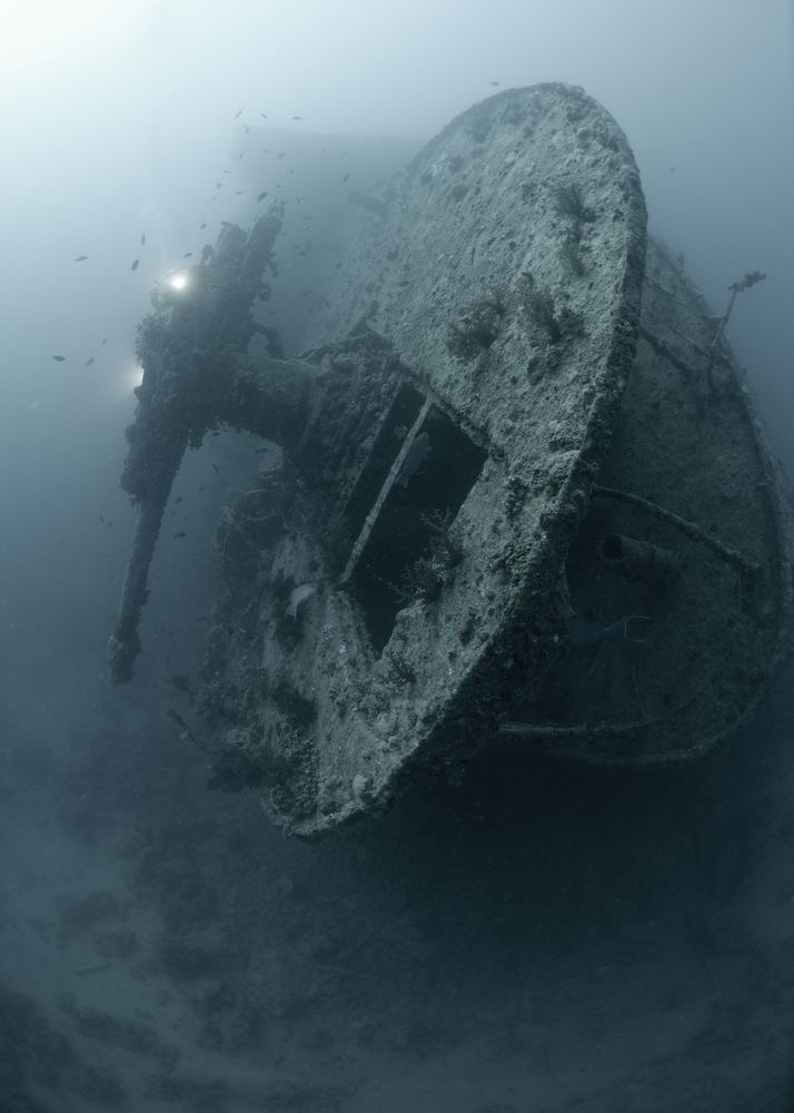 Divers approach the wreck of the SS Thistlegorm lying on its side in the murky waters of Egypt while fish swim towards her interior. 