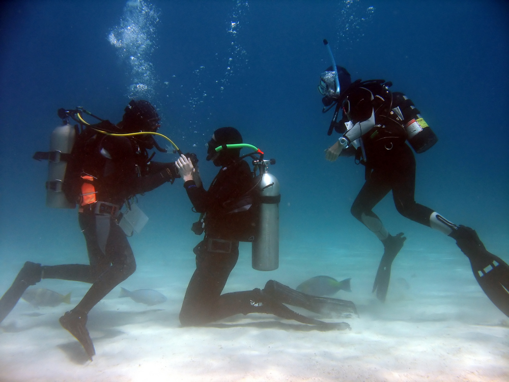 Bystander oversees two divers engage in buddy breathing on the sandy ocean bottom