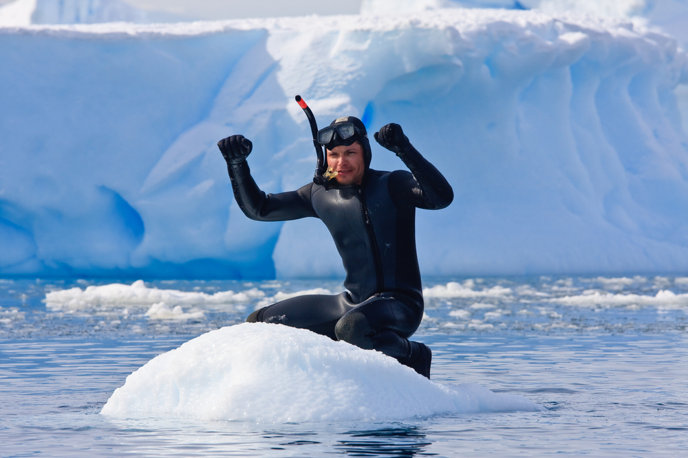 Ice diver showing off his ice diving gear including gloves while sitting atop an ice mound in the middle of the water