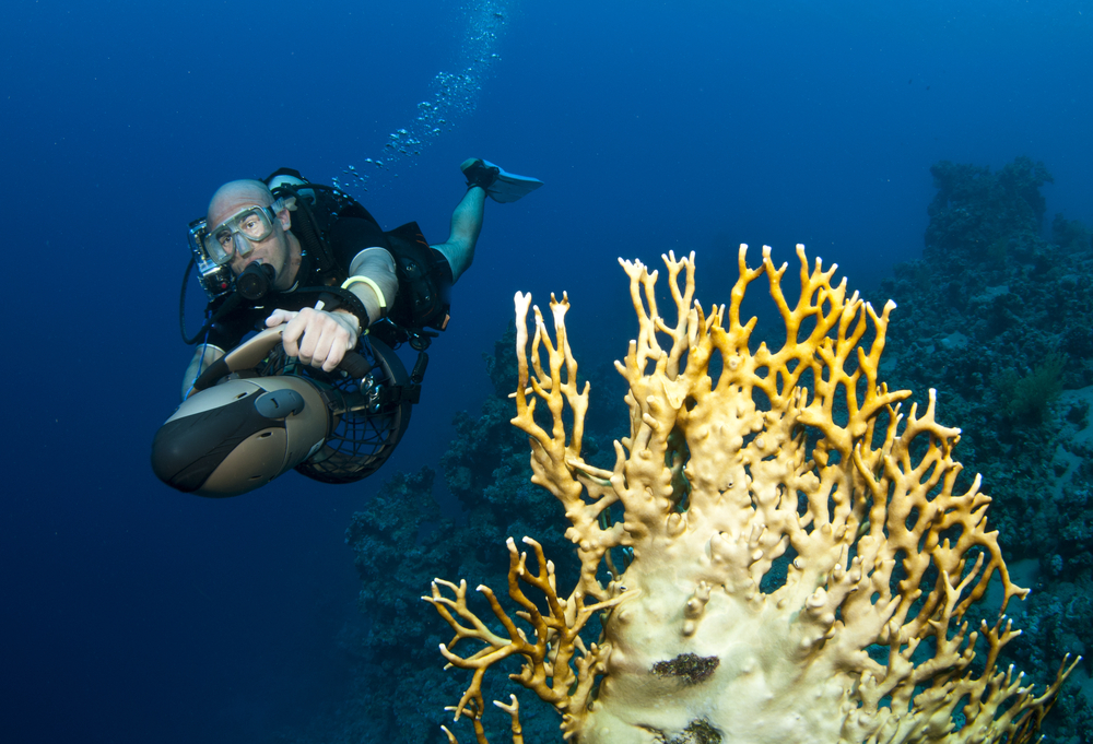 Male diver uses a diver propulsion vehicle (dpv) to help him explore more territory on his dive allowing him to observe the beautiful coral reefs and marine life with little exertion