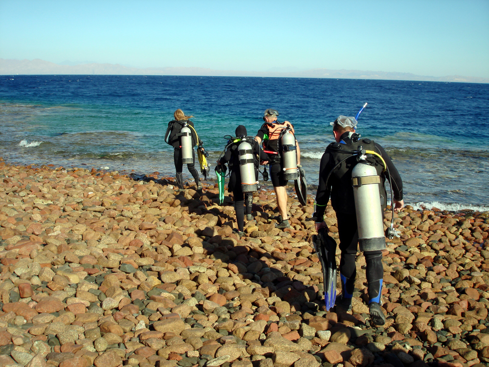 Group of shore divers in full kit walk across the rocky beaches towards the open ocean