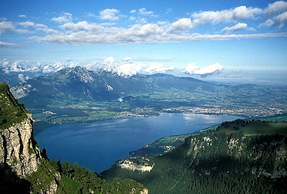 Aerial view of Lake Thun, also known as Thunersee, surrounded by lush greenery, mountains, and clouds in the backdrop