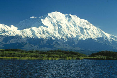Denali, also known as Mount McKinley, with its snow covered peaks surrounded by lush greenery and clear blue waters